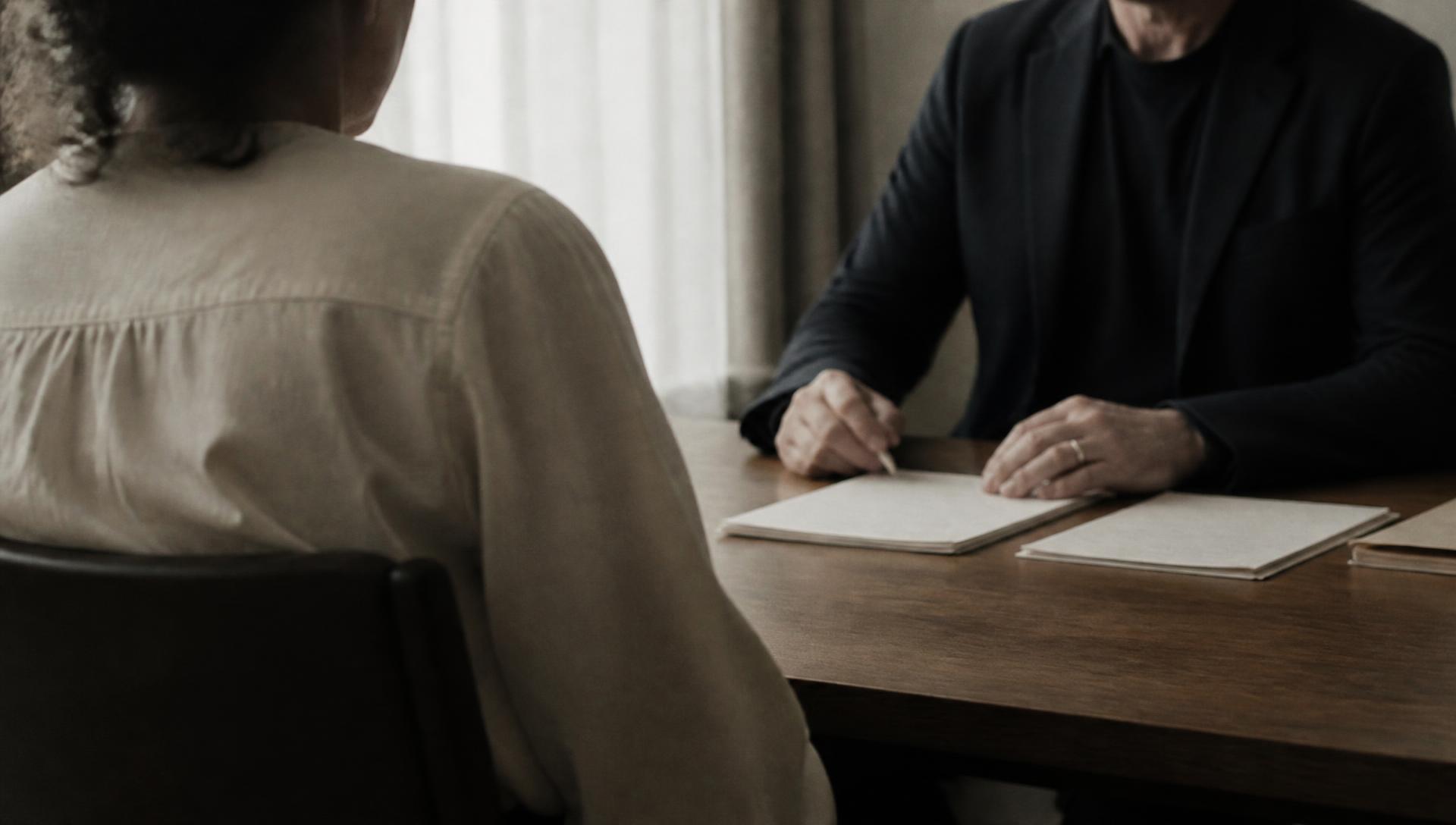 Two people seated across a table in a quiet meeting room, papers between them